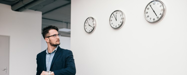 Hombre con traje mirando su reloj de pulsera junto a una pared con varios relojes, símbolo de la nueva ley de control horario